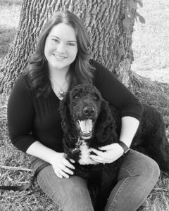 black and white photo of a smiling white woman sitting against a tree with a black Standard Poodle in her lap.