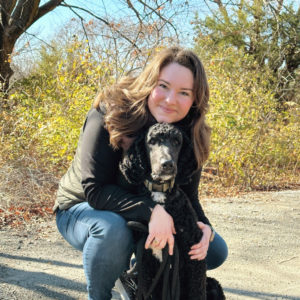 A white woman kneels down while outside on a gravel road with a black standard poodle between her knees.