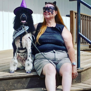 Black and white standard parti poodle with purple pointed hat and black with white bandana around her neck with purple nail polish on her nails while wearing a working harness. She is next to her team partner who has a black sleeveless shirt and grey shorts wearing a black mask on her face and black cat ears in her red hair.