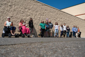 Outside in the sun against a beige rough stone wall, a group of 13 people, 9 service dogs, and two wheelchairs faces the camera.