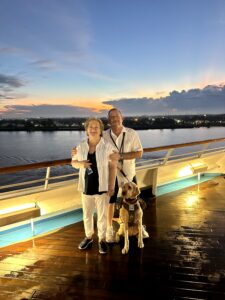 Photo 2: a tan Goldendoodle in a soft harness standing on the deck of the Carnival Ship Glory with his handler and the handler’s wife. The sunrise over New Orleans is in the background. The handler is wearing black shorts and a white top, while his wife is wearing white slacks, a blue shirt, and a white sleeved shirt over it.