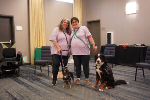 Two service dog teams pose for a picture in an open, carpeted convention meeting space. The people wear matching t-shirts, smiles, and pawprint stickers on their foreheads.
